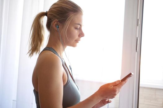 Young pretty sporty girl stands near the window and listens to music on headphones from smartphone. Sunny day