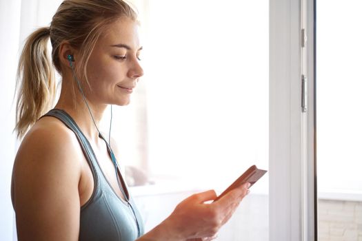 Happy young pretty sporty girl stands near the window and listens to music on headphones from smartphone