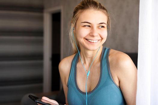 Young pretty sporty girl at home listens to music on headphones from smartphone