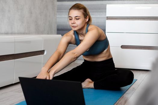 Pretty young woman in sportswear watching online video on laptop and doing exercises at home. Distant training with personal trainer, online education