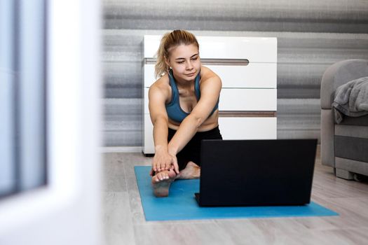 Pretty young woman in sportswear watching online video on laptop and doing exercises at home. Distant training with personal trainer, online education