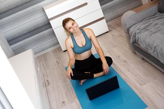 Happy cheery young fitness woman sit on blue yoga mat at home near laptop