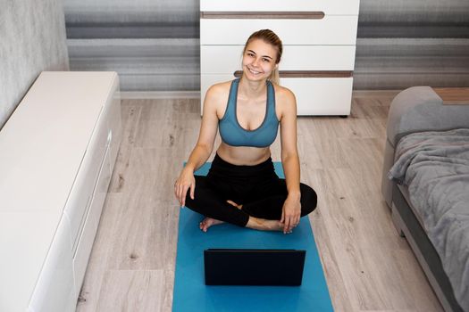 Happy cheery young fitness woman sit on blue yoga mat at home near laptop