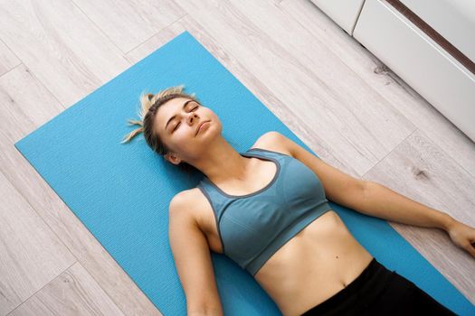 Top view of beautiful young woman lying on yoga mat after workout. Fit female relaxing on floor at home.