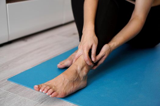 Young woman suffering from pain in ankle or foot injury while sitting on stretching mat. Healthcare and lifestyle concept