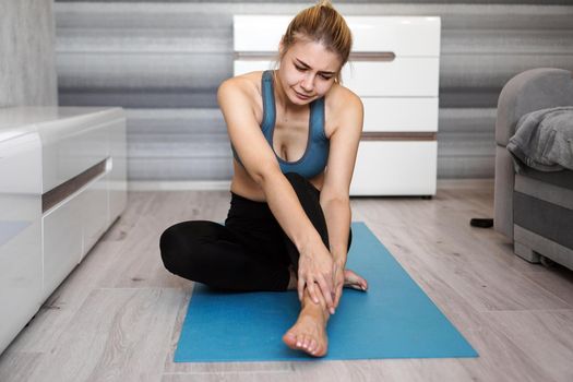 Unhappy woman sitting on the yoga mat with ankle injury, feeling pain. Health care and execise concept.