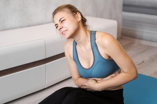 Portrait of unhappy young woman sitting on yoga mat, touching her stomach after training, suffering from stomach, feeling pain, side view