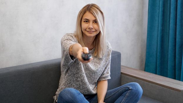 Young woman switches the television channels with the remote control. She smiles and looks at the camera