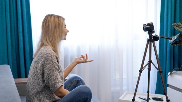 Young woman in casual clothes records herself on camera on tripod. Side view. Cheerful female blogger recording video at home