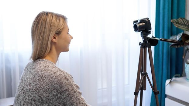 Young woman in casual clothes records herself on camera on tripod. Side view. Cheerful female blogger recording video at home