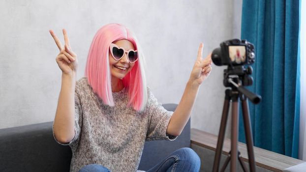 Happy teen girl blogger with smiling face in pink wigs and glasses. Shows the victory sign, looking at camera recording live vlog, making videocall