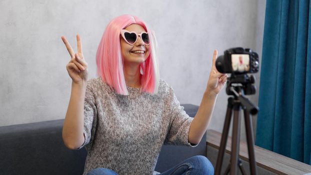 Happy teen girl blogger with smiling face in pink wig and glasses. Shows the victory sign, looking at camera recording live vlog