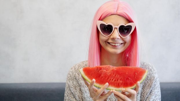Young woman with piece of watermelon in hand, adorable female smiling at camera. Woman with pink wig and heart-shaped glasses. Place for text