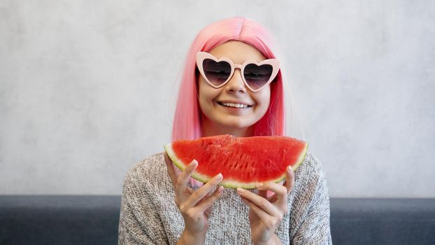 Indoor shot of young woman with piece of watermelon in hand, adorable female smiling at camera. Woman wears pink wig and heart-shaped glasses