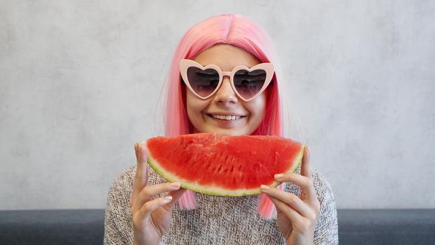 Indoor shot of young woman with piece of watermelon in hand, adorable female smiling at camera. Woman wears pink wig and heart-shaped glasses