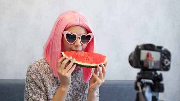 Positive blogger records a video about healthy eating and holds a watermelon in her hands. Woman wearing pink wig and heart shaped glasses