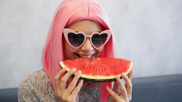 Indoor shot of young woman with piece of watermelon in hand, adorable female smiling at camera. Woman wears pink wig and heart-shaped glasses