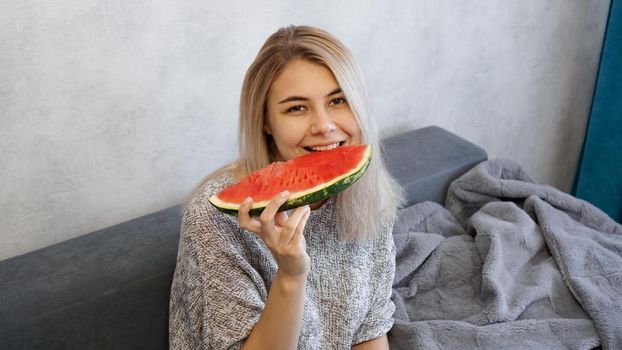 Young attractive woman bites a piece of watermelon. Woman at home in a cozy interior looking at camera and smiling