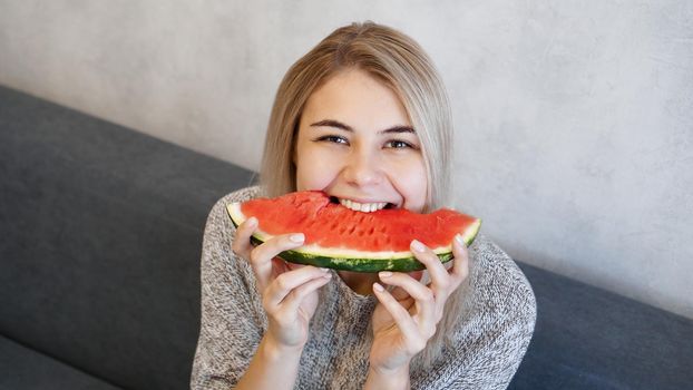 Young attractive woman bites a piece of watermelon. Woman at home in a cozy interior looking at camera and smiling