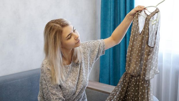 Pretty woman holds a hanger with a dress and looks at it. Woman chooses outfit at home