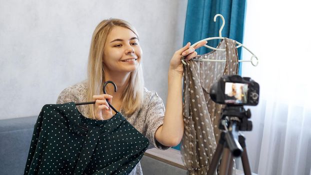 Fashion blogger choosing her outfit, holding two dresses on hangers and recording video for blog. Woman in front of the camera