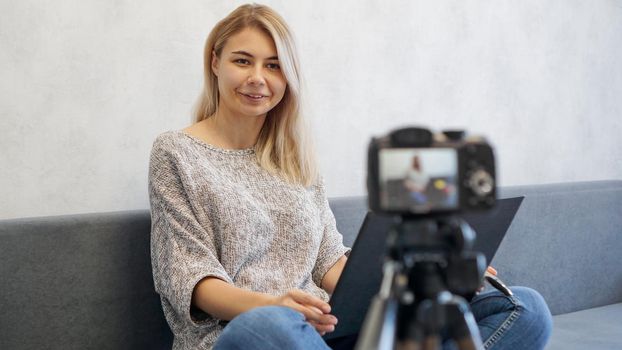 Young female blogger with laptop looking at camera. She talks about business or records a lecture for students. She is positive and smiling.