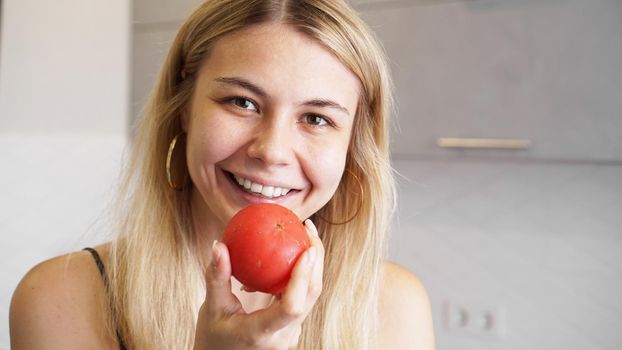 Young happy woman holding tomato and smiling on background of modern white kitchen. Healthy food concept. Home cooking