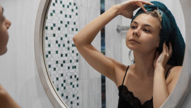 Woman with a towel on hair looks in the mirror in the bath room