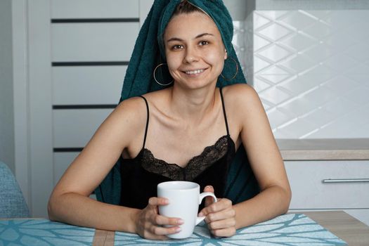 Beautiful girl in a towel on hair sits in the kitchen with a cup of coffee. Woman looking at camera and smiling