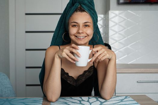 Beautiful girl in a towel on hair sits in the kitchen with a cup of coffee. Woman looking at camera and smiling