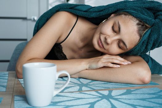 Tired woman sleeping on the table. Beautiful girl in a towel on hair sits with a cup of coffee. Lazy sleepy morning