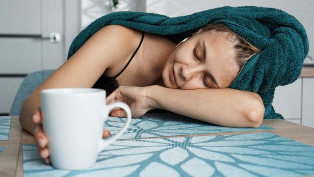Tired woman sleeping on the table. Beautiful woman in a towel on hair. Hand reaches for the mug. Lazy sleepy morning