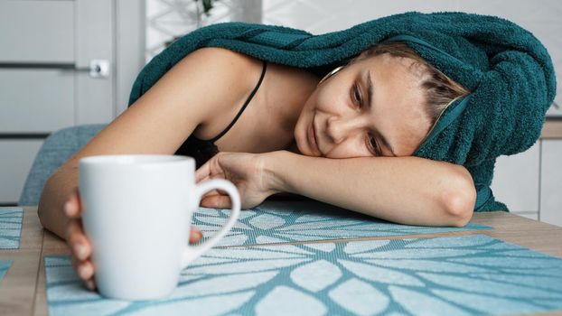 Tired woman sleeping on the table. Beautiful woman in a towel on hair. Hand reaches for the mug. Lazy sleepy morning