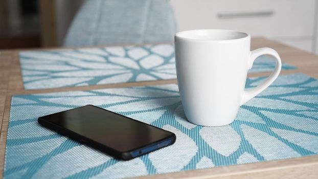 Clean white mug with handle and smart phone on blue table behind blurred kitchen background