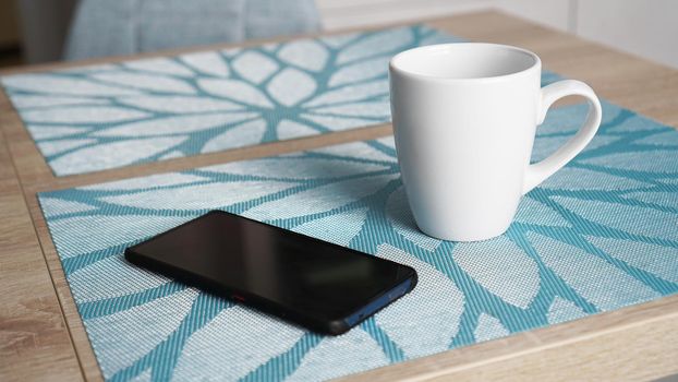 Clean white mug with handle and smart phone on blue table behind blurred kitchen background