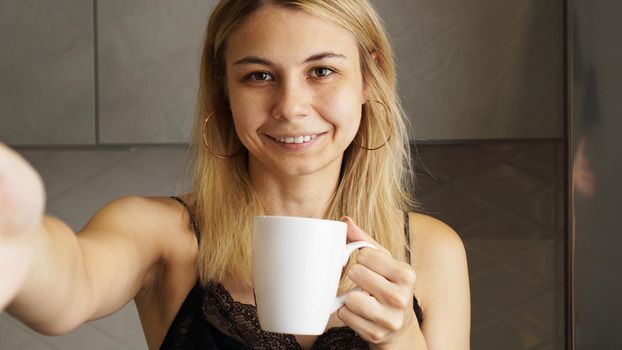 Selfie portrait of attractive young woman with cup of tea smiling to camera in kitchen of modern apartment