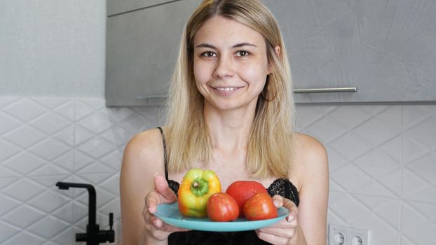 Woman holding plate with fresh vegetables and smiling on a background of kitchen