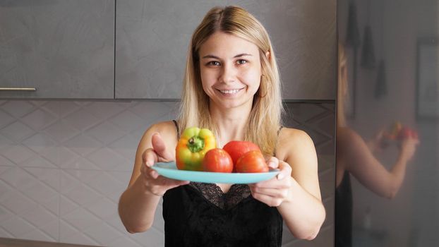 Woman holding plate with fresh vegetables and smiling on a background of kitchen