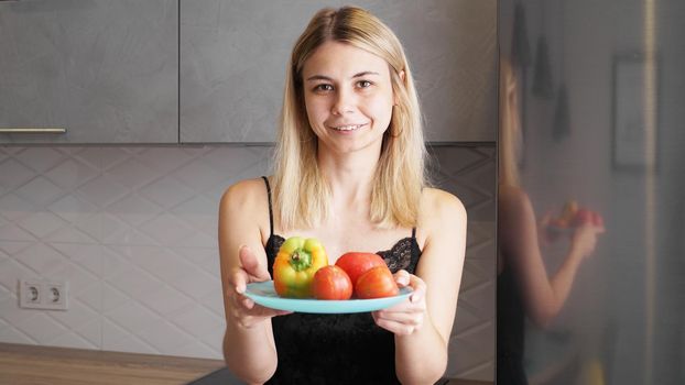 Woman holding plate with fresh vegetables and smiling on a background of kitchen
