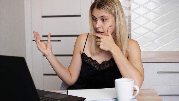 Portrait of an attractive woman at the table with cup and laptop. Confused young woman having problem with computer, looking at laptop screen