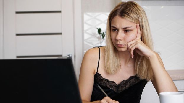 Portrait of an attractive woman at the table with cup and laptop. Confused young woman having problem with computer, looking at laptop screen