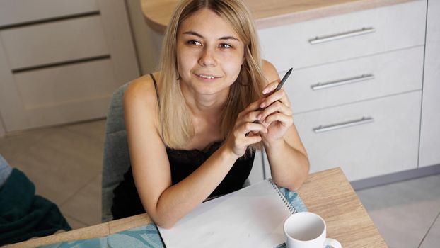 A woman sits in a bright kitchen and writes a letter or her wishes. A girl dreams. A cup of coffee is on the table.
