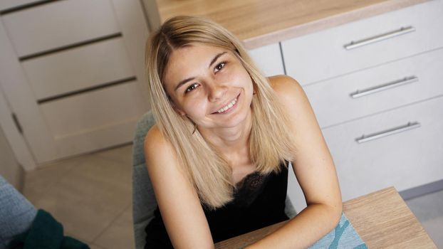 Cheerful young female sitting at table in cozy styled room while smiling and looking at camera