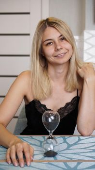 Woman looks at camers and smiles, time concept. She in a bright kitchen and waits. Hourglass on the table