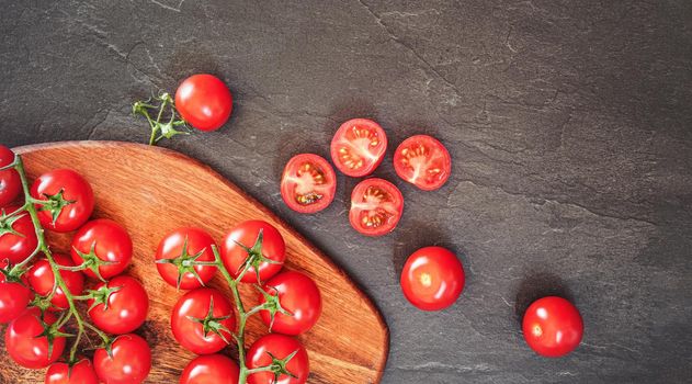 Fresh red cherry tomatoes on wooden cutting board, some sliced to half over black slate like board near.