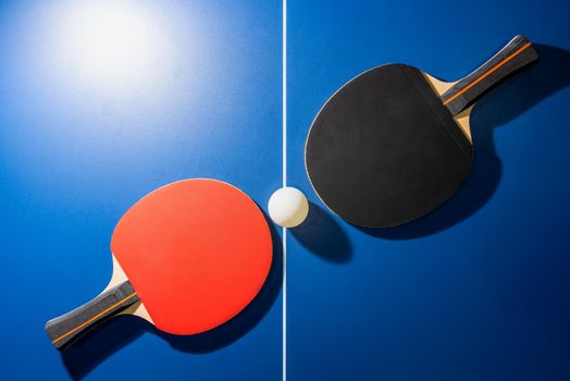 Top view black and red table tennis racket and a white ping pong ball on the blue ping pong table with a bright spotlight, Two table tennis paddle is a sports competition equipment for indoor exercise