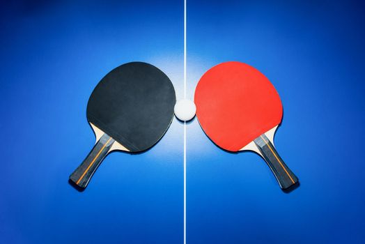 Top view black and red table tennis racket and a white ping pong ball on the blue ping pong table with a bright spotlight, Two table tennis paddle is a sports competition equipment for indoor exercise