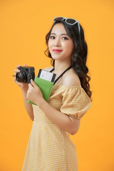 Image of happy asian young woman tourist standing isolated over yellow  background holding camera and passport with tickets.