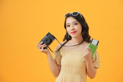 Portrait of a happy young woman holding camera and showing passport while standing isolated over yellow background