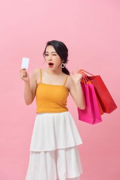 Full length portrait of a happy beautiful girl wearing dress and holding shopping bags and showing credit card isolated over pink background